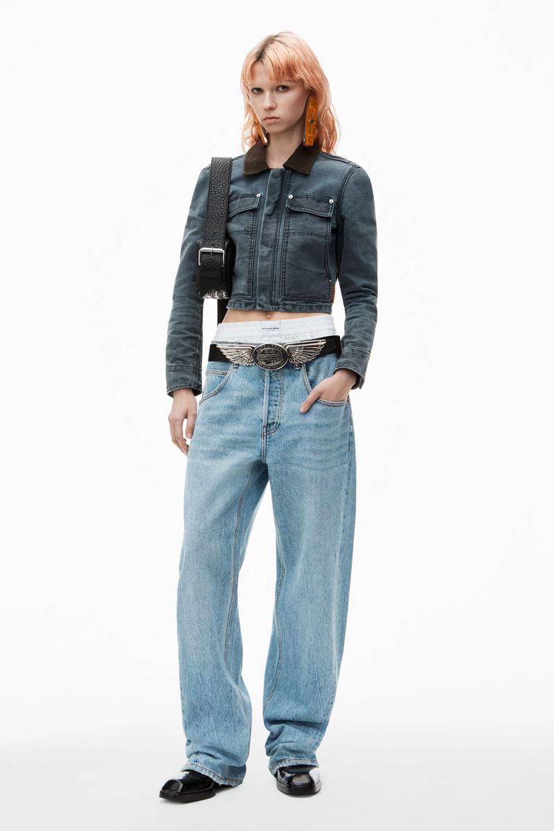 A woman with peach-colored hair poses in a cropped denim jacket, oversized jeans, and black shoes against a stark white background.
