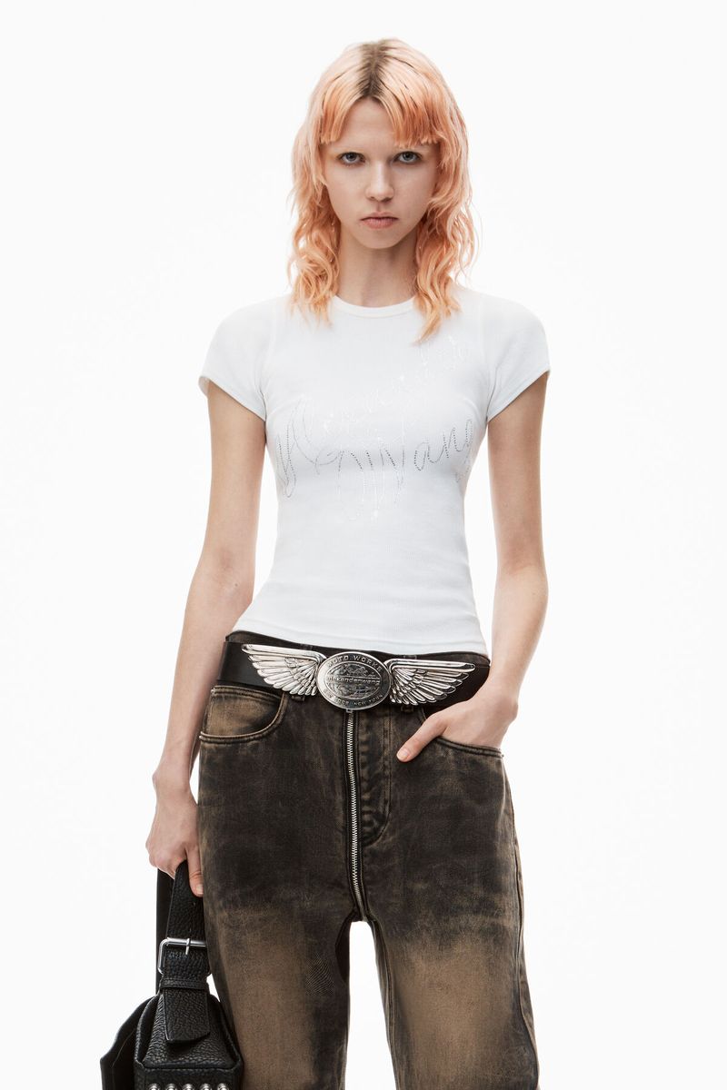 A young adult woman poses indoors in a white T-shirt, distressed denim jeans, and a decorative wing-buckle belt, holding a small bag.