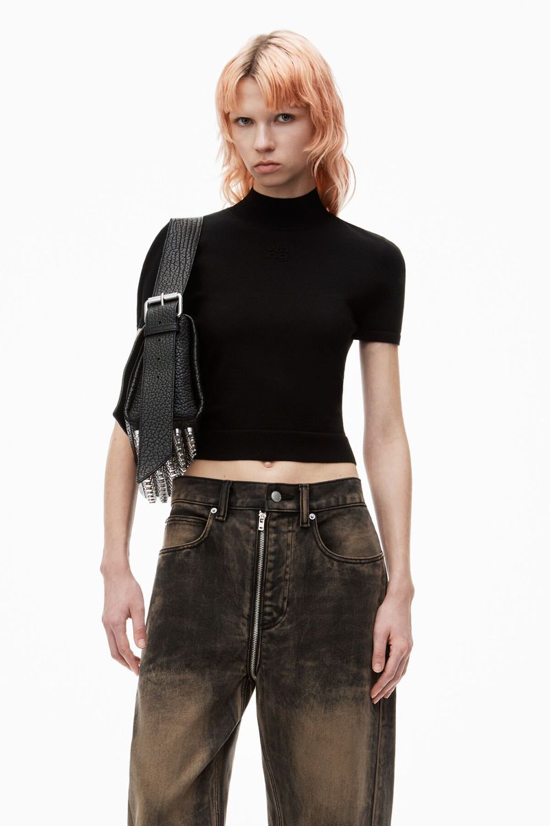 Woman with pink hair wears a black top, distressed denim pants, and a black shoulder bag in a studio shot against a white backdrop.
