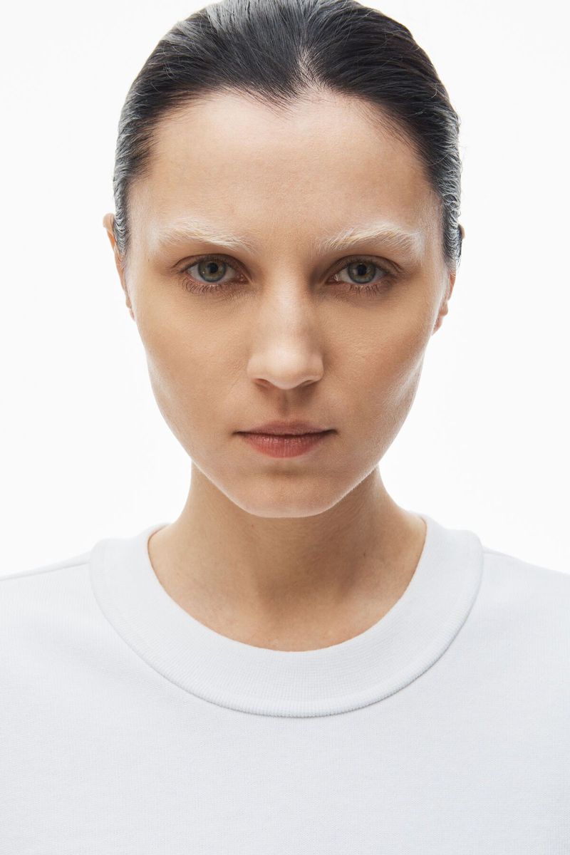 Close-up portrait of a young woman with fair skin, bleached eyebrows, wearing a white t-shirt, against a bright white background.