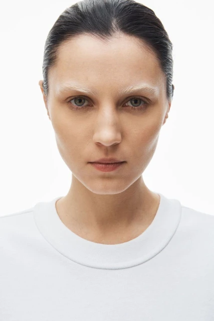 Close-up portrait of a young woman with fair skin, bleached eyebrows, wearing a white t-shirt, against a bright white background.