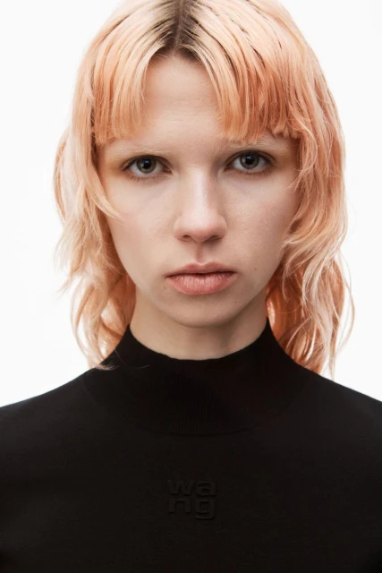Close-up portrait of a blonde adult woman with fair skin, wearing a black turtleneck sweater, set against a bright white background.