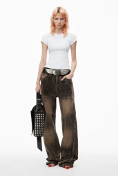 Model with reddish-blonde hair wearing a white t-shirt and distressed baggy jeans holding a studded black handbag against a plain white background
