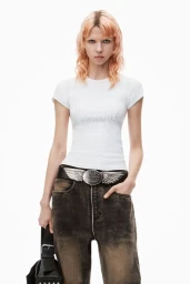 Model with peach hair wearing a white t-shirt, distressed denim jeans, and a winged belt against a white background