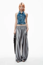 Model wearing a denim halter vest and glossy grey trousers holding a black strap against a white background