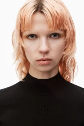 Close-up portrait of a model with pink-toned hair wearing a black mock-neck top against a seamless white background.