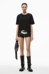 Model standing against a white backdrop wearing a black t-shirt, leather shorts, and boots while holding a studded bag.