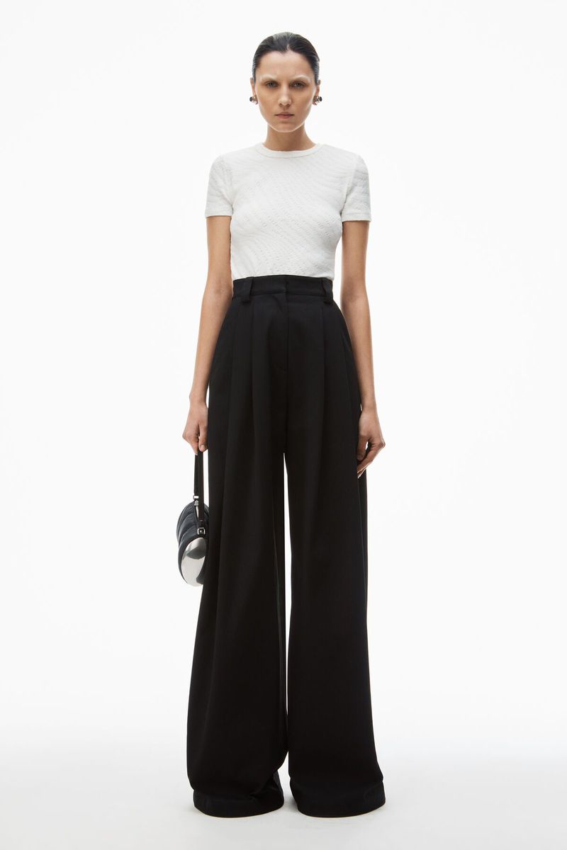 Model wearing a white t-shirt and wide black trousers holding a small black handbag against a white background