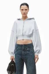 Model wearing a light grey cropped zip hoodie and dark denim jeans holding a black studded bag against a white background.