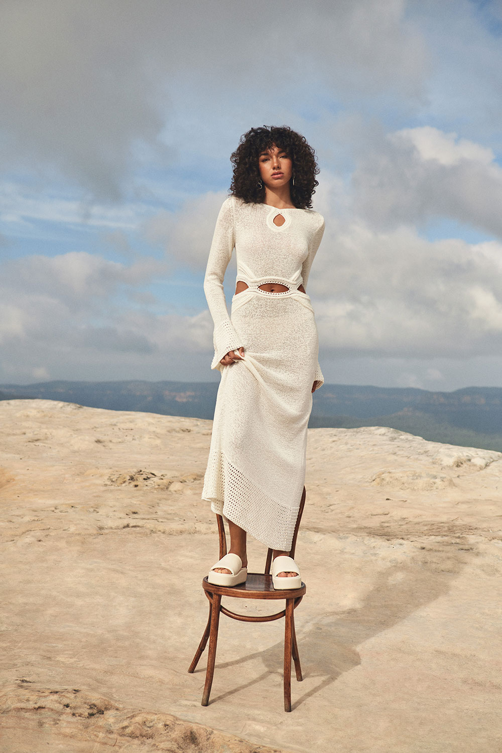 An adult female stands on a wooden chair outdoors in a long-sleeved cream dress, accessorized with jewelry and platform sandals in a desert setting.