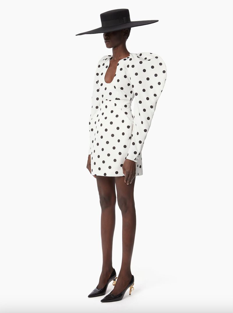A single Black woman stands in a white polka dot dress and a wide-brimmed black hat against a clean white backdrop in a fashion setting.