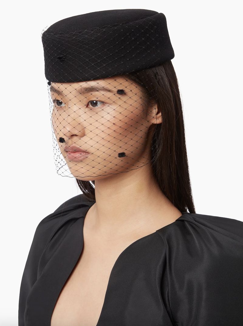 Asian woman wears a black pillbox hat with a black veil, posing in a studio with a bright background; fashion portrait.