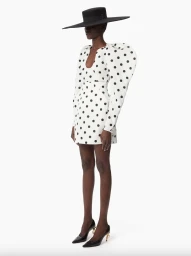 Model wearing a white polka dot mini dress with exaggerated shoulders and wide-brim black hat on white seamless background.