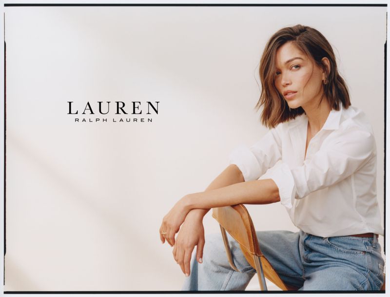 A brunette woman in a white shirt and blue jeans is seated on a wood chair in a fashion shot with Ralph Lauren branding.