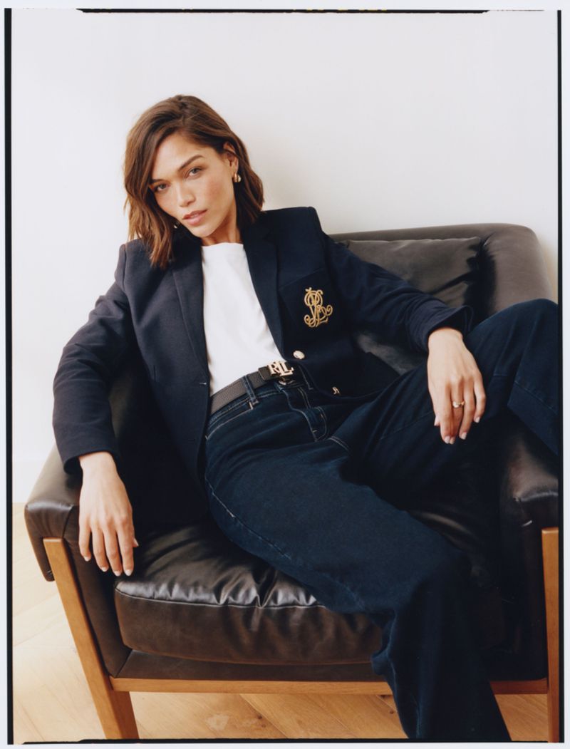 Adult woman with short brunette hair sits in an armchair wearing a navy blazer, white t-shirt, dark wash jeans, and a black belt.