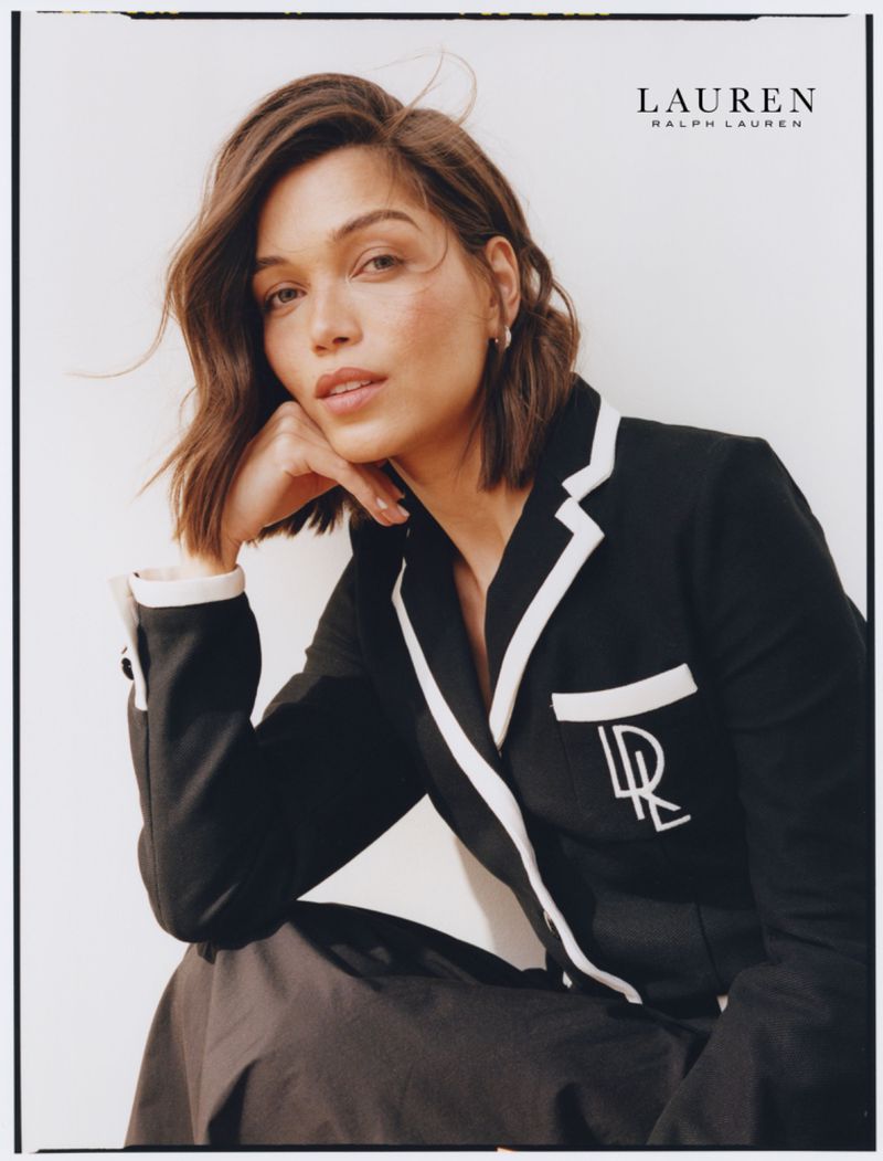 Close-up shot of an adult woman with short brown hair in a black jacket, posing in front of a neutral background for a fashion advertisement.