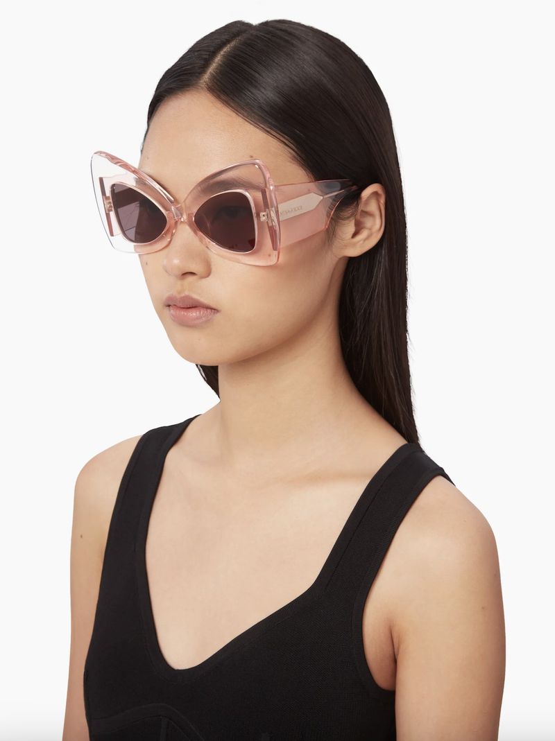 Portrait of a woman wearing oversized butterfly sunglasses with a black tank top against a white backdrop in a brightly lit studio.