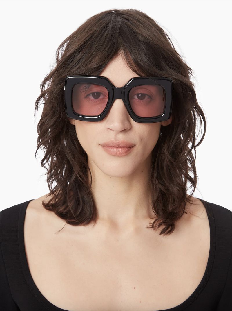 A woman with fair skin and dark wavy hair wears large, square black sunglasses with pink lenses in a studio headshot.