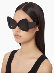 Model wearing bold black winged sunglasses and a black sleeveless top against a clean white background