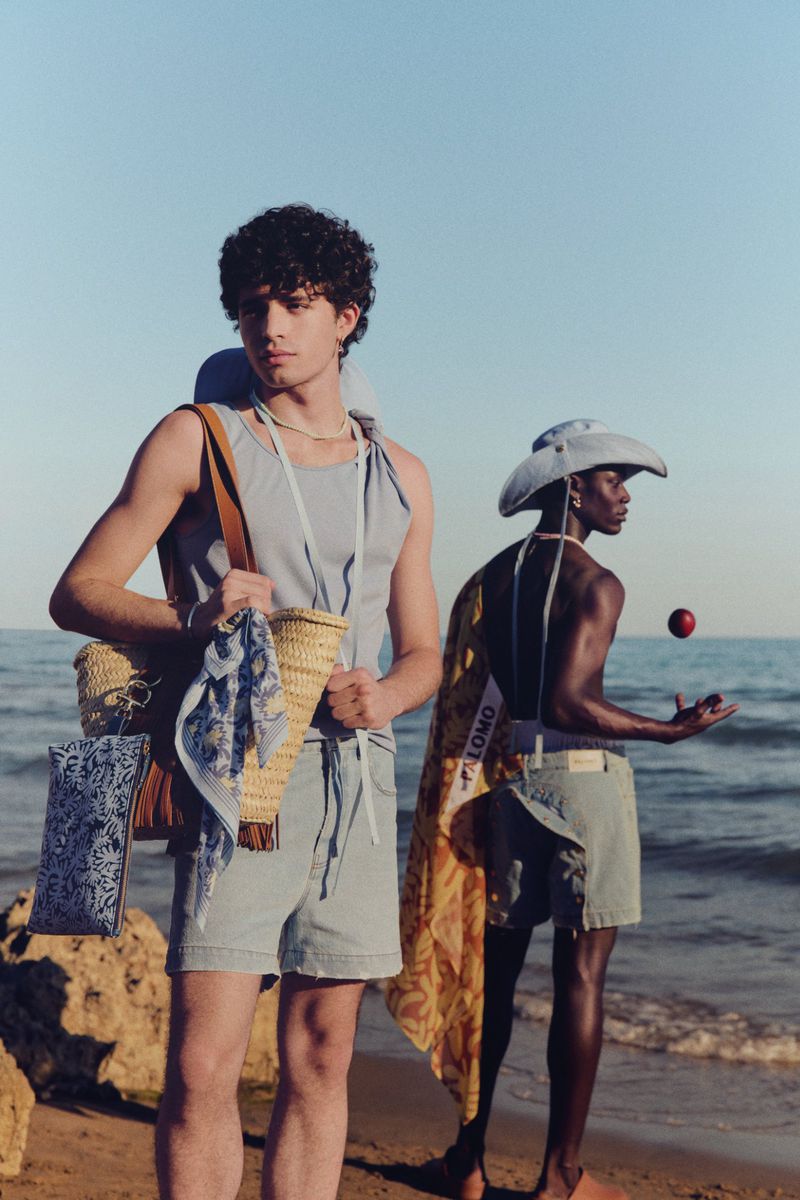 Two men pose on a beach with straw bags, light blue outfits, and neutral tones under bright skies, showcasing summer fashion.