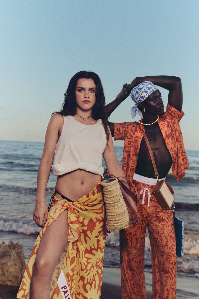 Two models pose on a beach at sunset. One woman wears a white tank top and orange sarong, the other wears an orange patterned shirt and pants.