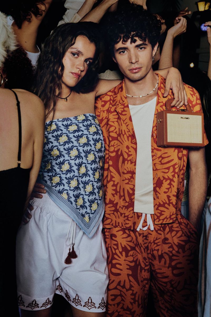 Two people in patterned summer outfits with a brown handbag, posing in front of a blurred, lively crowd indoors with dark lighting.