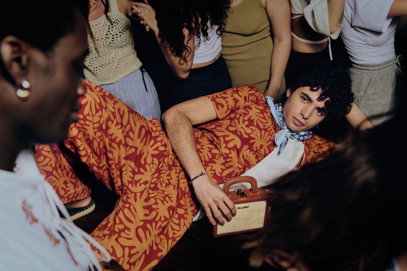 A young man in a patterned orange suit poses with a bag in a crowded indoor setting, surrounded by other people and soft, ambient lighting.
