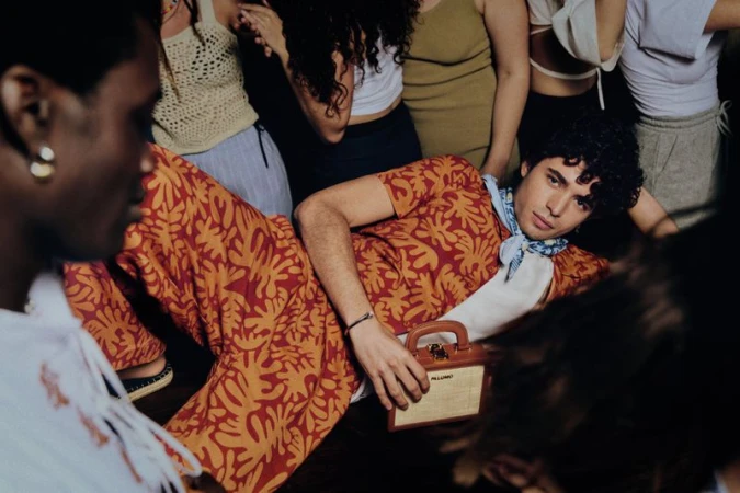 A young man in a patterned orange suit poses with a bag in a crowded indoor setting, surrounded by other people and soft, ambient lighting.