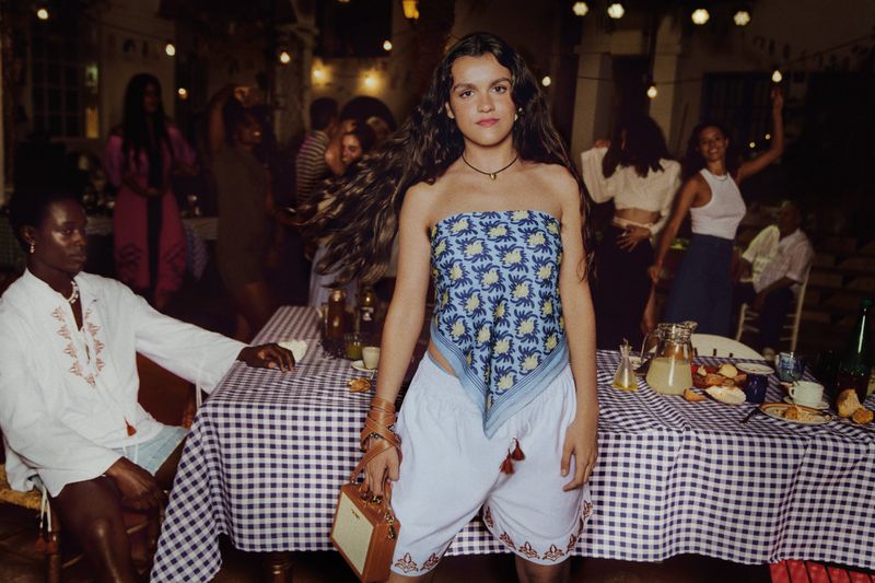 Young woman in patterned scarf top stands at party with food-laden table and dancing group in background, illuminated by string lights.