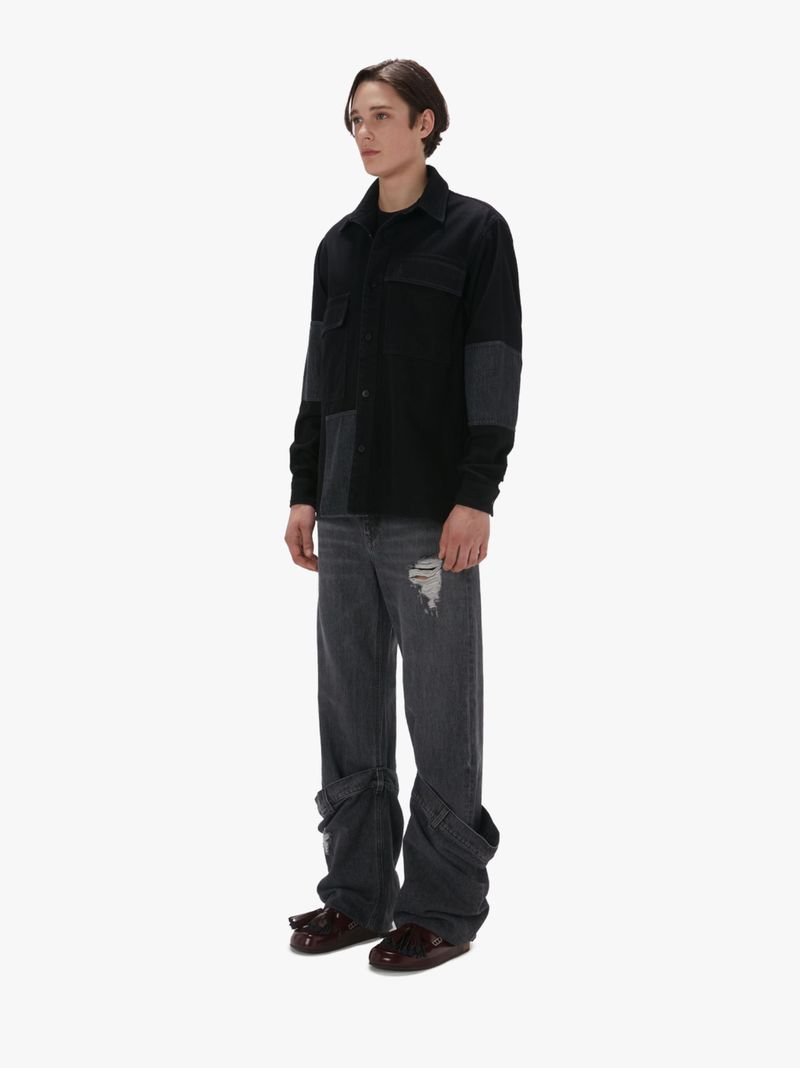 Full-length shot of a young adult male in a black patchwork shirt and grey distressed jeans against a white background in a studio.