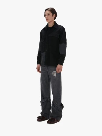 Full-length shot of a young adult male in a black patchwork shirt and grey distressed jeans against a white background in a studio.