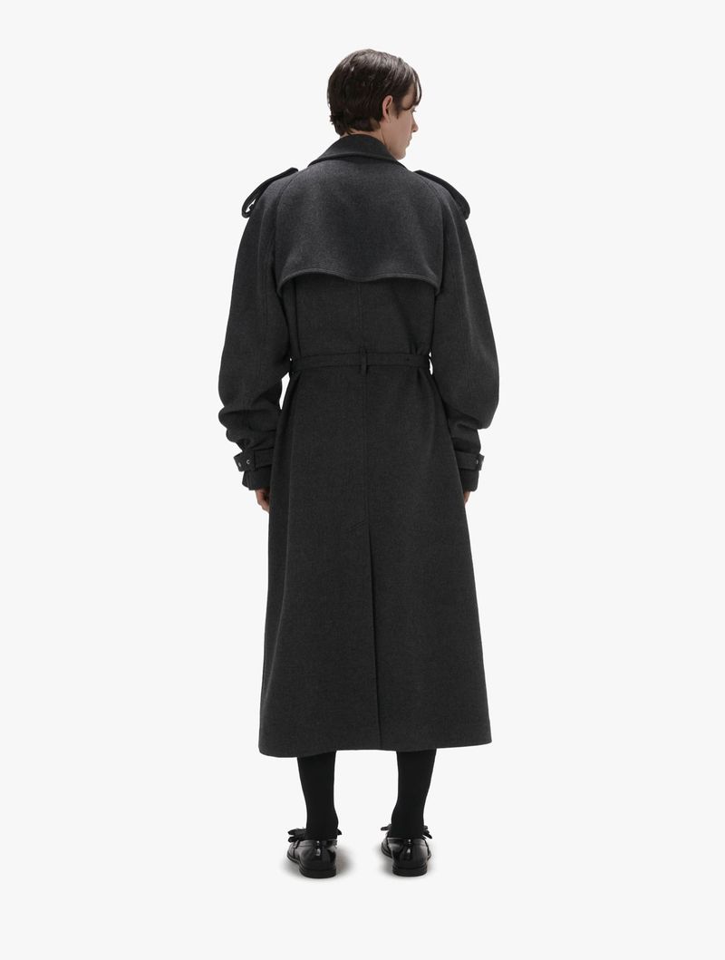 Back view of a person wearing an oversized grey trench coat, black socks, and black Mary Jane shoes against a white backdrop.