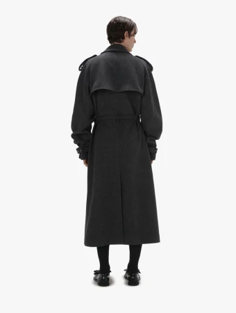 Back view of a person wearing an oversized grey trench coat, black socks, and black Mary Jane shoes against a white backdrop.