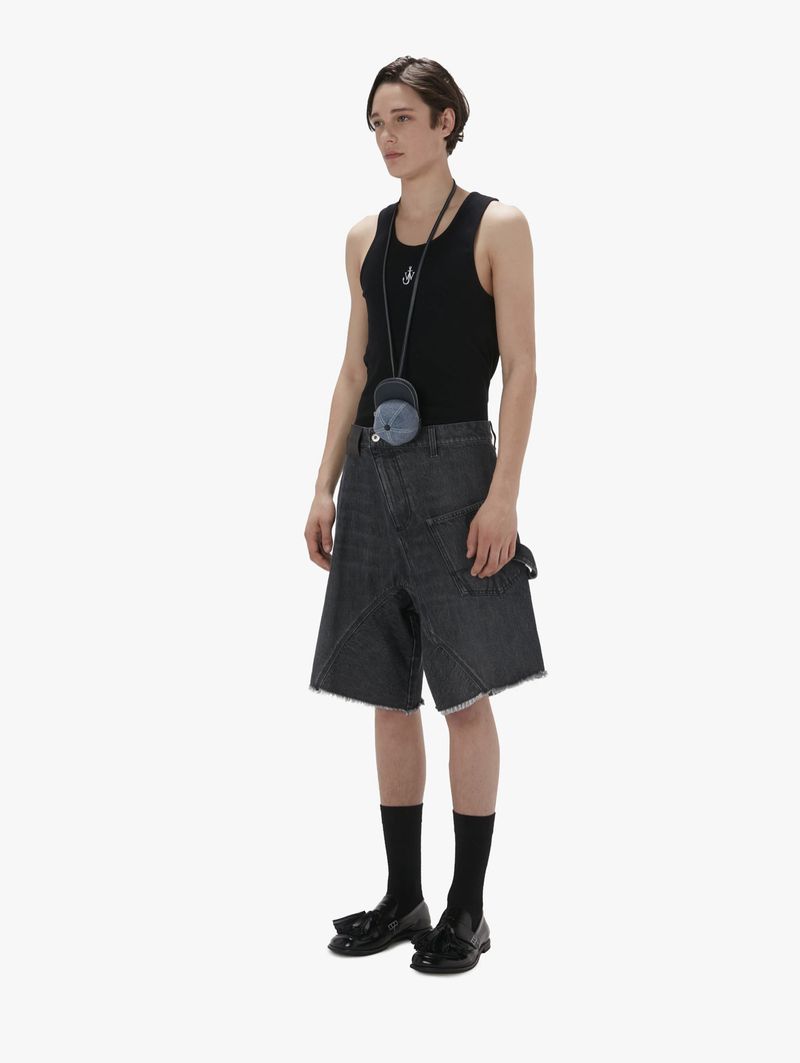 A slender man poses in a studio wearing a black tank top, denim shorts, black socks, and black shoes against a stark white background.