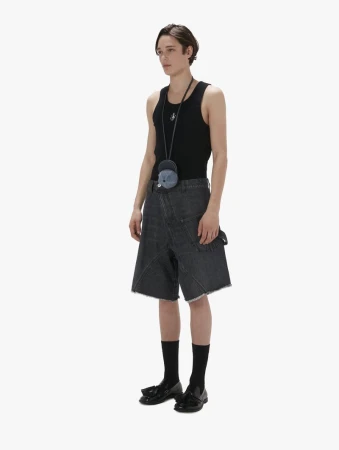 A slender man poses in a studio wearing a black tank top, denim shorts, black socks, and black shoes against a stark white background.