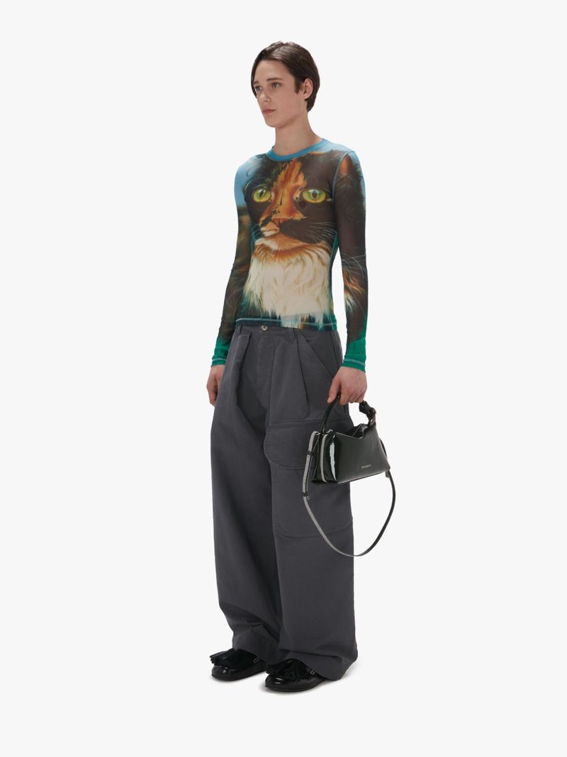 Fashion shot of a person wearing a cat print top, grey trousers, and black shoes, standing in a bright photo studio with a handbag.
