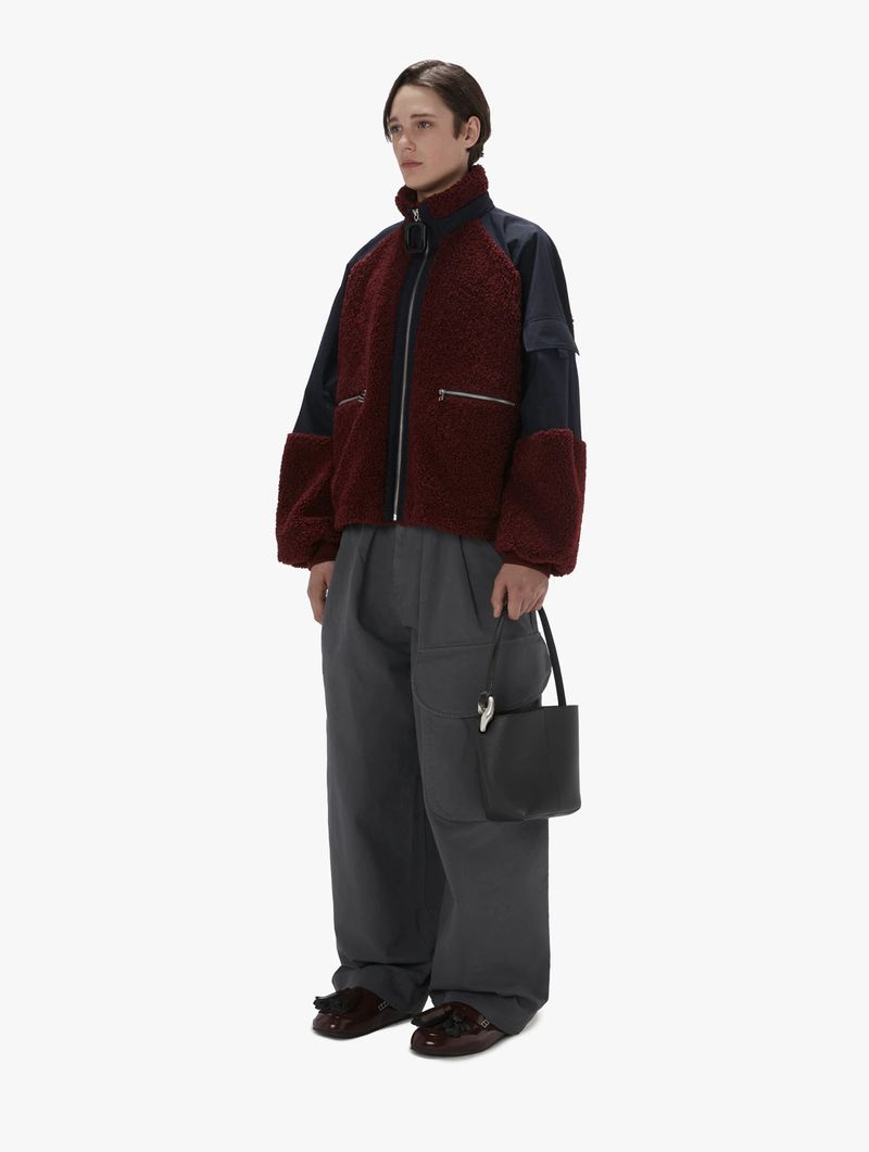 A young adult male stands in a brightly lit studio wearing a two-tone sherpa jacket, loose grey trousers, brown loafers, and a black purse.