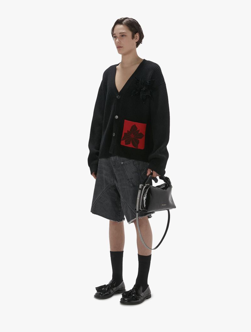 Full-length shot of an adult male wearing a black patterned cardigan with grey denim shorts, black socks, and black loafers, holding a black purse.
