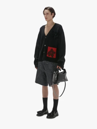 Full-length shot of an adult male wearing a black patterned cardigan with grey denim shorts, black socks, and black loafers, holding a black purse.