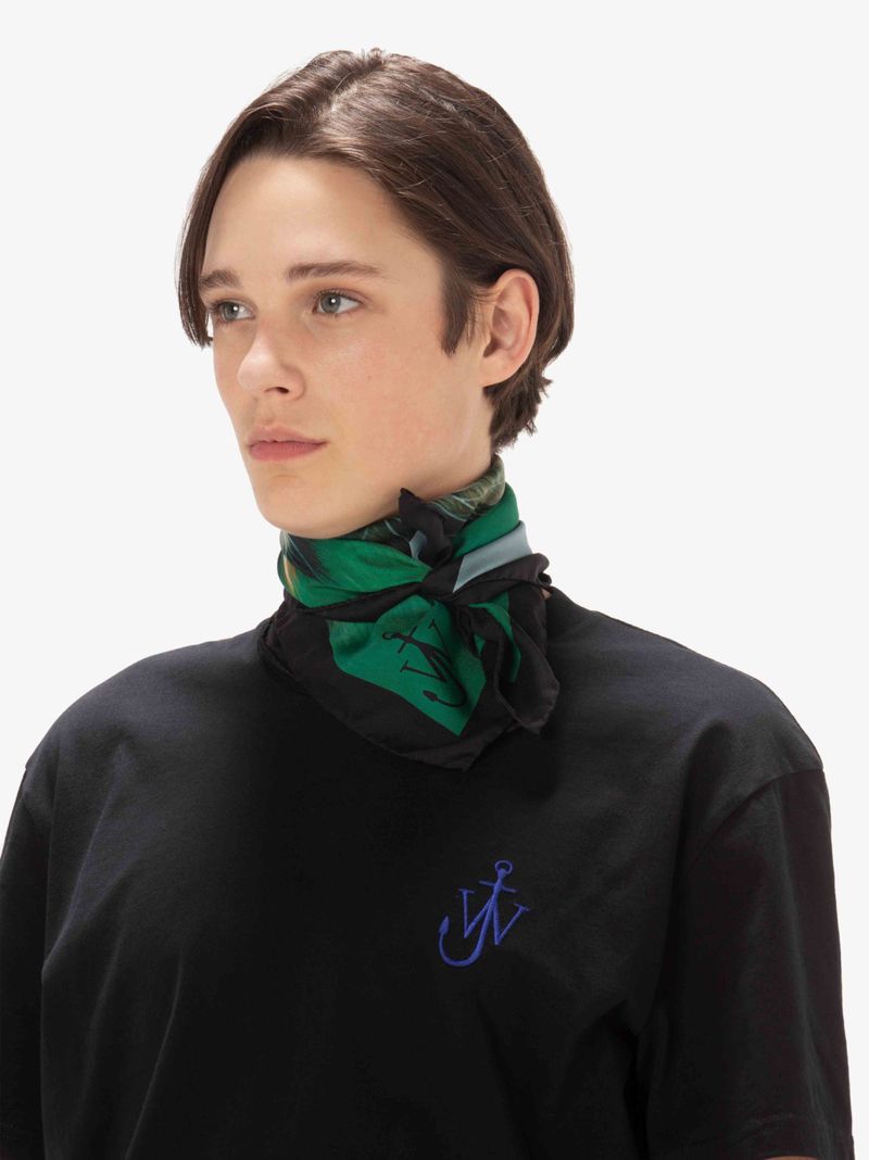 Young adult male in a black t-shirt with a blue logo and a green-and-black printed scarf tied around his neck against a white backdrop.