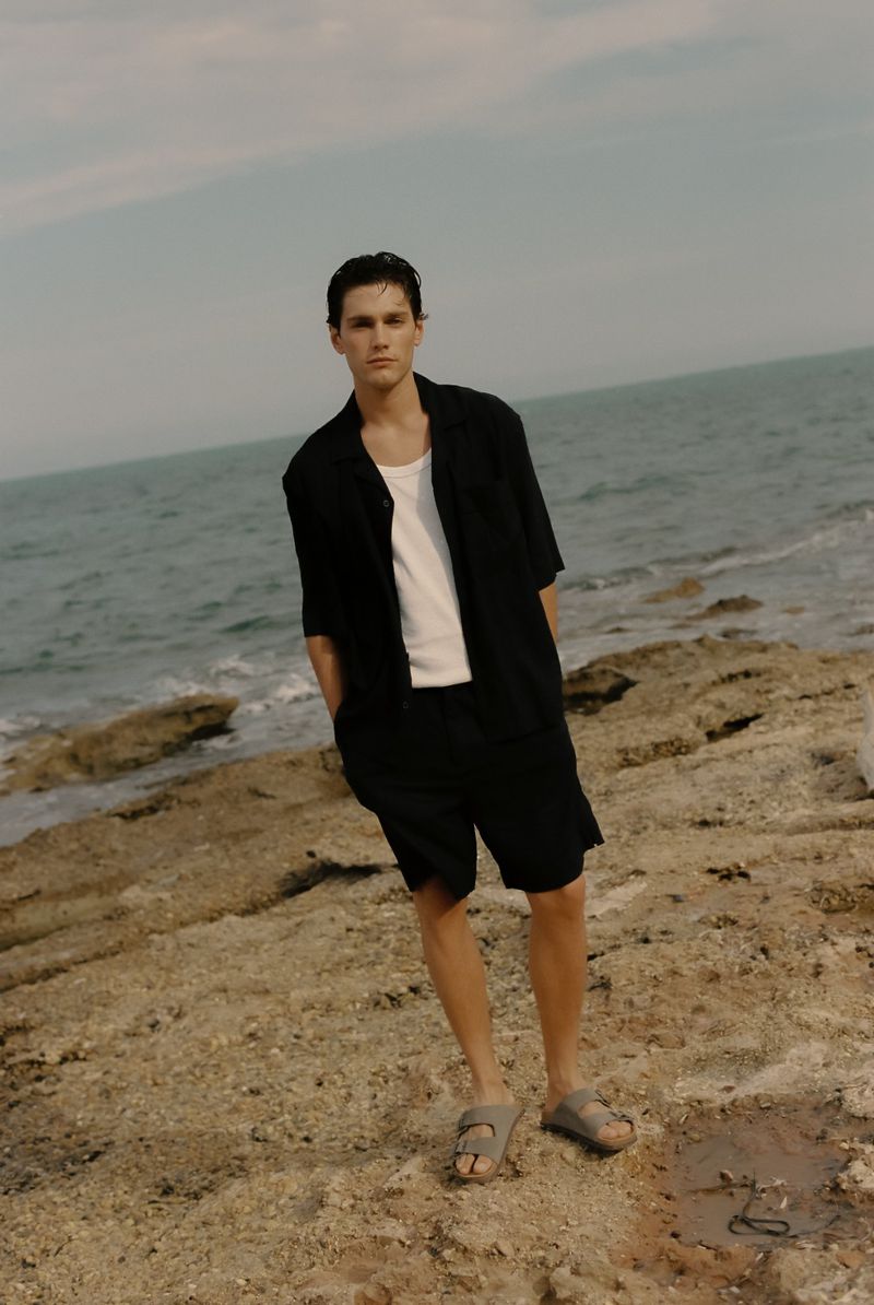 An adult male stands on rocks near the ocean, wearing a dark collared shirt, white t-shirt, dark shorts, and sandals, with a cloudy sky.