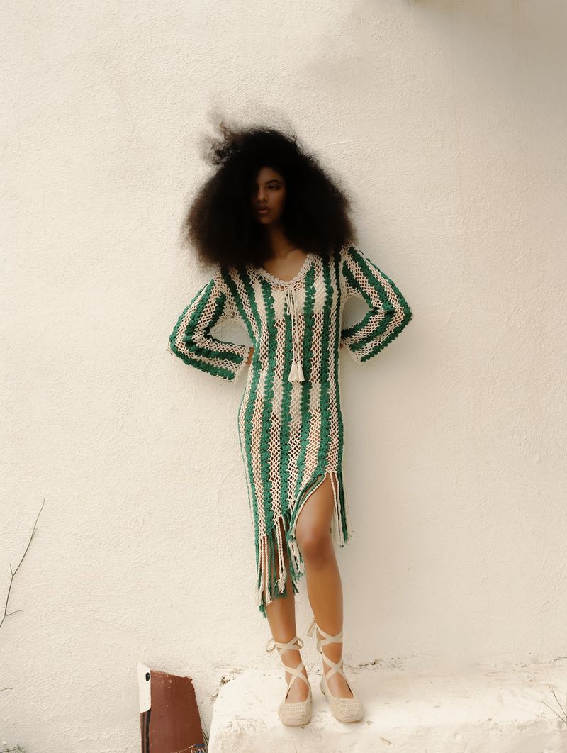 A curly-haired model stands on a white step in a green-and-cream striped crochet dress with gladiator-style cream-colored espadrille sandals and a white wall background.