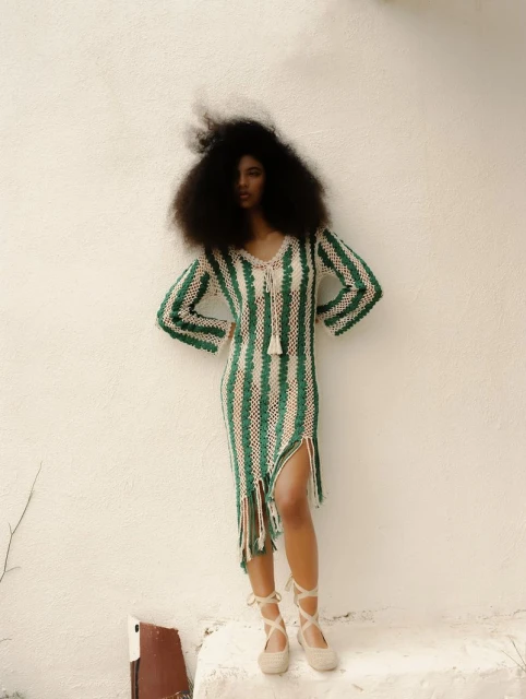 A curly-haired model stands on a white step in a green-and-cream striped crochet dress with gladiator-style cream-colored espadrille sandals and a white wall background.
