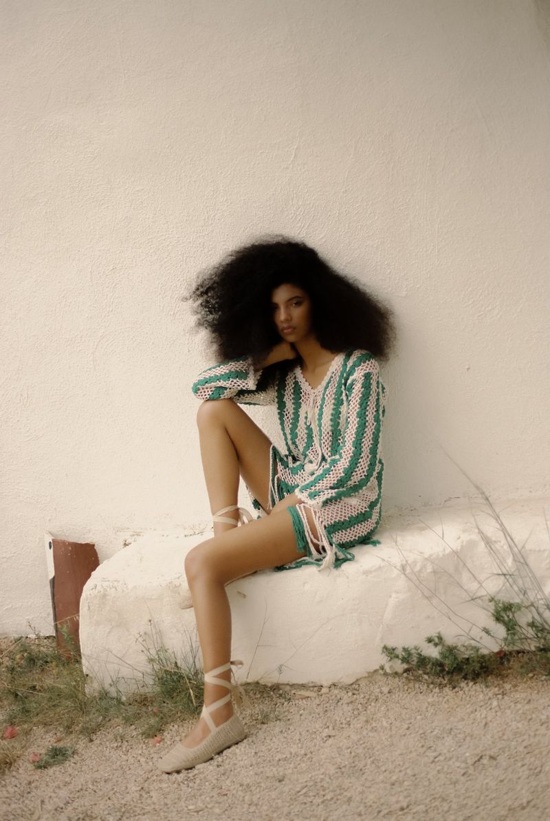 A woman with afro hairstyle sits on a concrete block outside, wearing a pink and green striped crochet mini dress and ballet shoes.