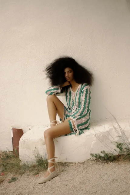 A woman with afro hairstyle sits on a concrete block outside, wearing a pink and green striped crochet mini dress and ballet shoes.