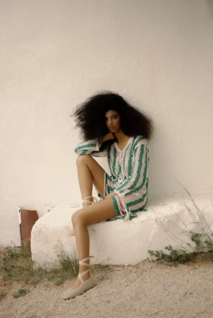 A woman with afro hairstyle sits on a concrete block outside, wearing a pink and green striped crochet mini dress and ballet shoes.