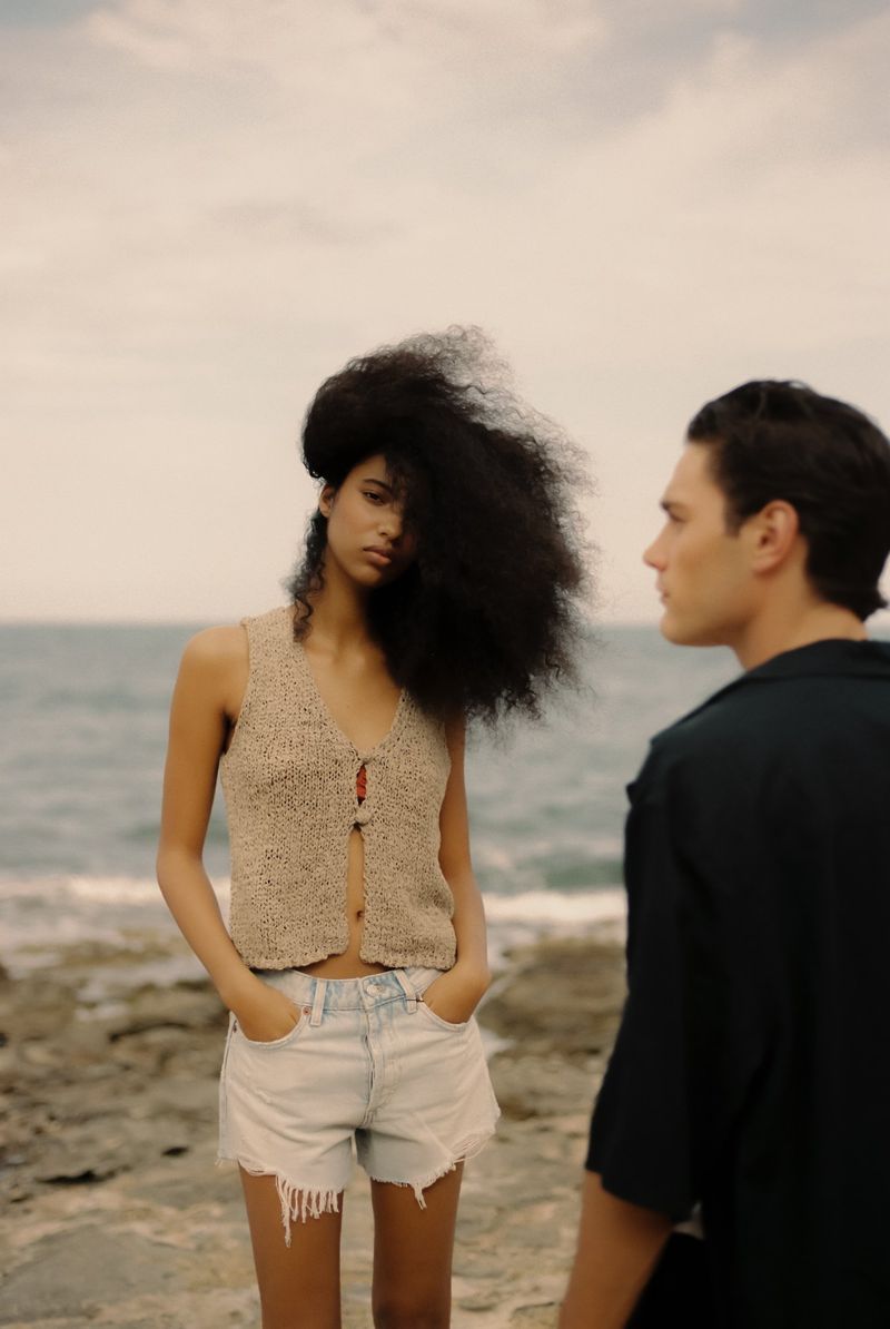 A woman in a crochet top and denim shorts stands on rocks by the ocean, with a man looking toward her in a serene outdoor setting.