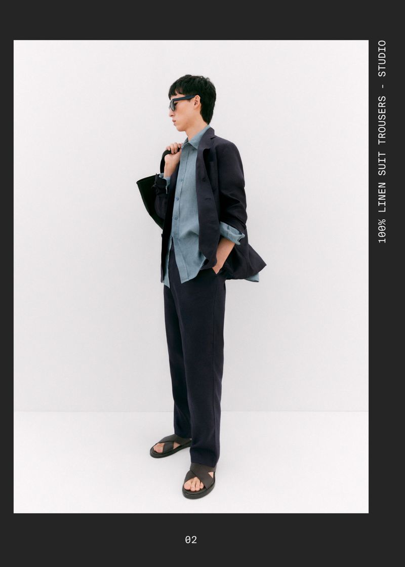 A stylish male fashion model poses indoors, wearing a blue linen shirt, suit trousers, jacket, sandals, and sunglasses against a white backdrop.