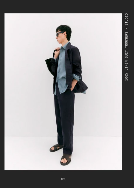 A stylish male fashion model poses indoors, wearing a blue linen shirt, suit trousers, jacket, sandals, and sunglasses against a white backdrop.