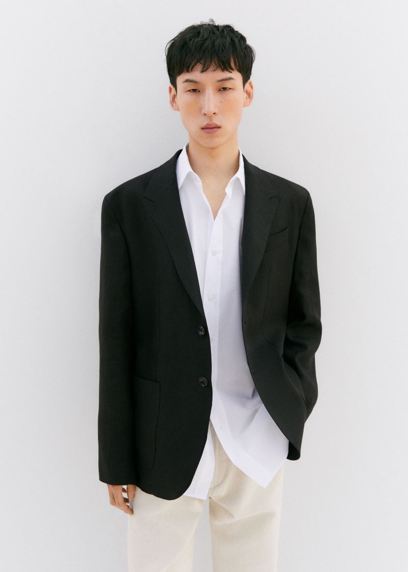 Man in a black blazer and white shirt poses in front of a white wall, conveying a modern, minimalist fashion aesthetic.